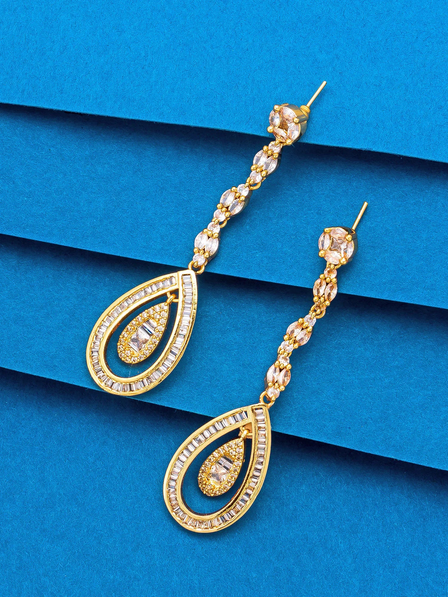 A pair of gold teardrop dangle earrings with clear rhinestone accents, displayed on a textured blue background.