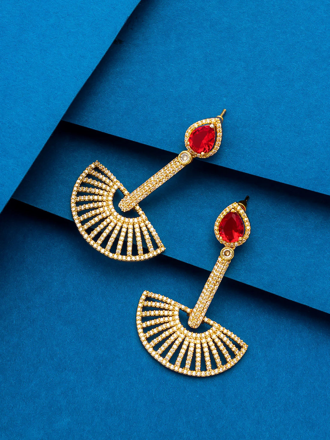 A pair of gold-toned dangle earrings with teardrop-shaped red gemstones and fan-shaped pavé-set details, displayed on a textured blue background.