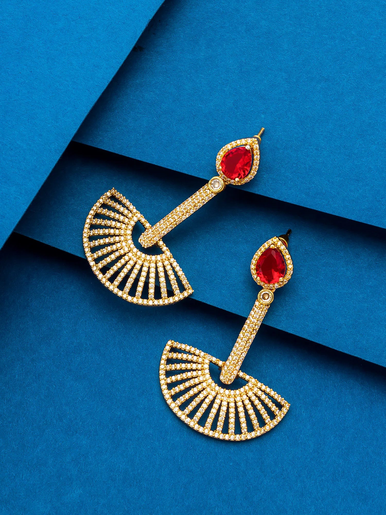 A pair of gold-toned dangle earrings with teardrop-shaped red gemstones and fan-shaped pavé-set details, displayed on a textured blue background.