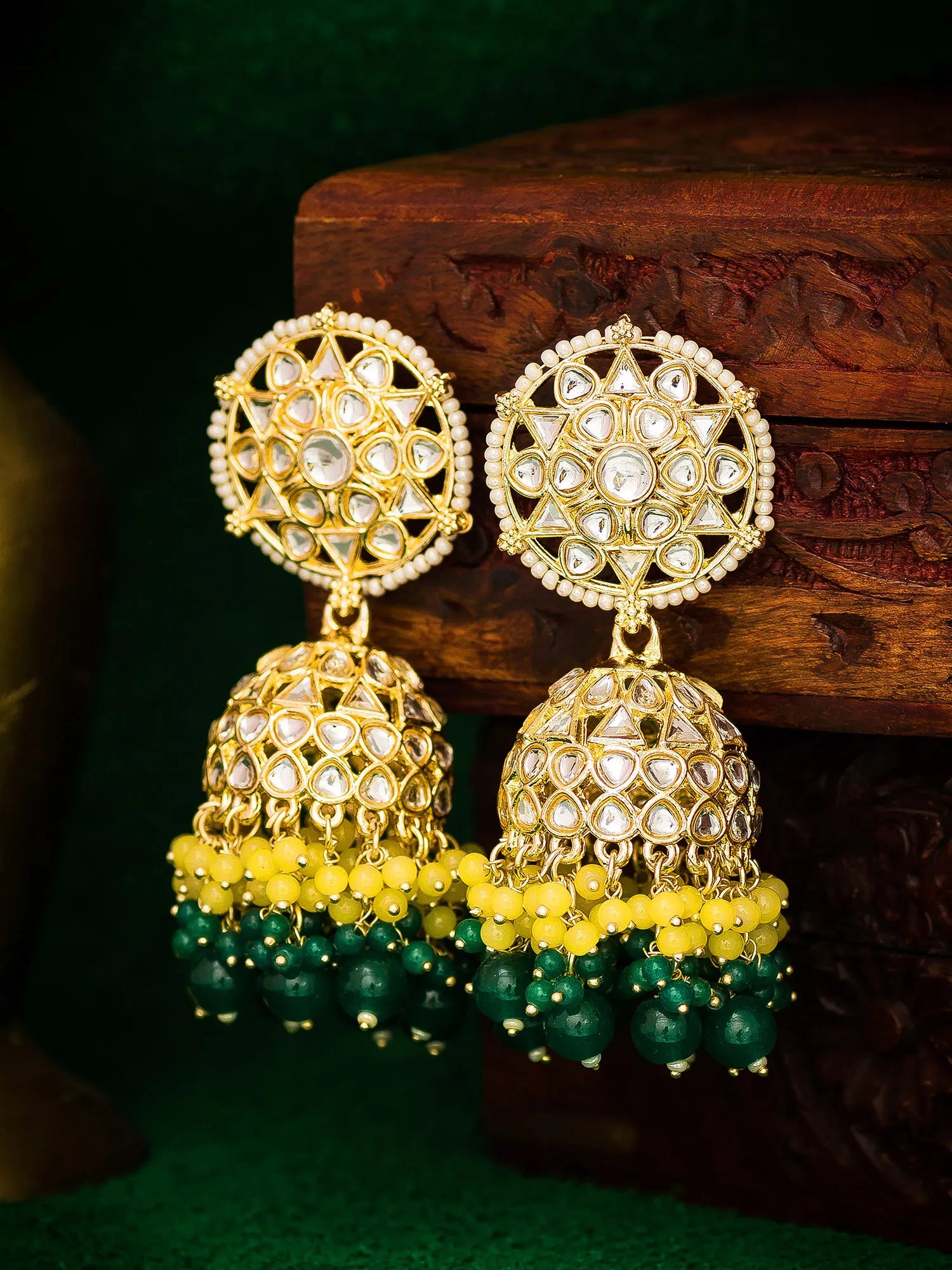 Gold-plated Indian jhumka earrings with white stones, yellow beads, and green bead drops, displayed on a carved wooden block.