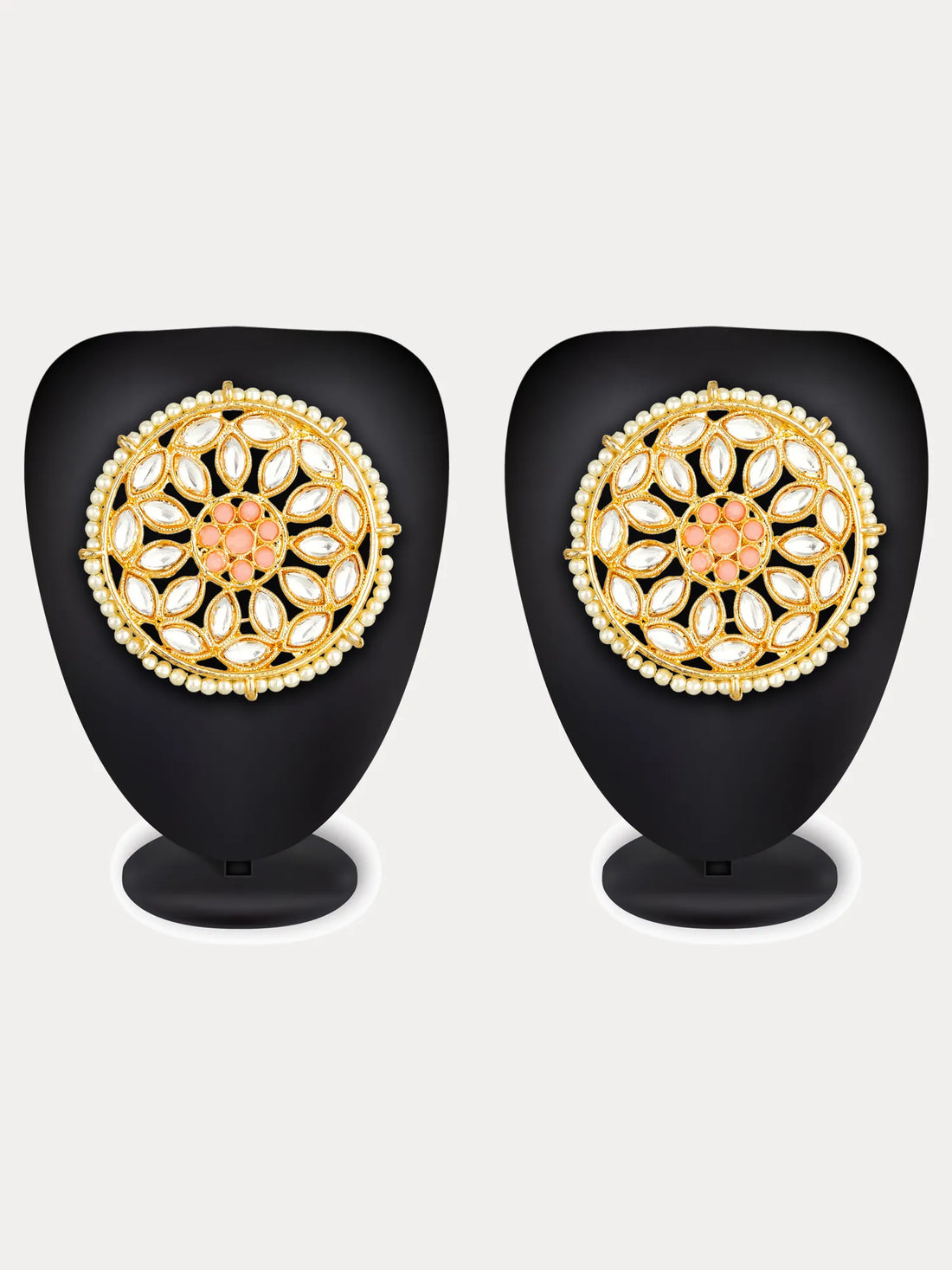 A pair of ornate gold-toned Indian jhumka earrings decorated with white stones and a coral-pink floral center, displayed on a carved dark wood surface with a draped red fabric background.