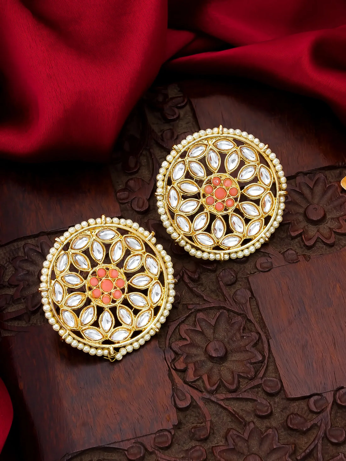 A pair of ornate gold-toned Indian jhumka earrings decorated with white stones and a coral-pink floral center, displayed on a carved dark wood surface with a draped red fabric background.