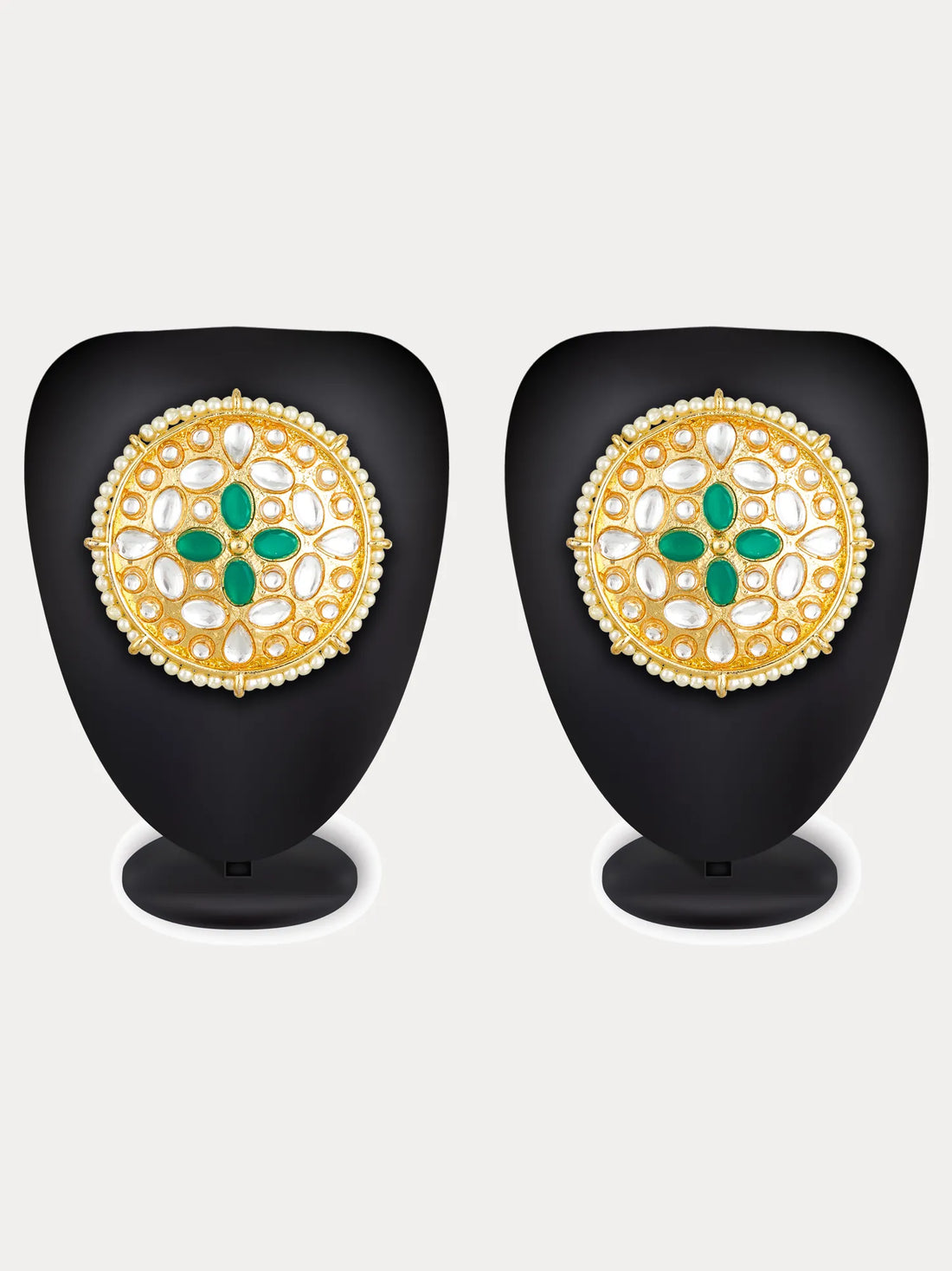 A pair of ornate Indian gold earrings decorated with white stones, green enamel, and a border of small white pearls, displayed on a carved wooden surface with a red satin cloth background.