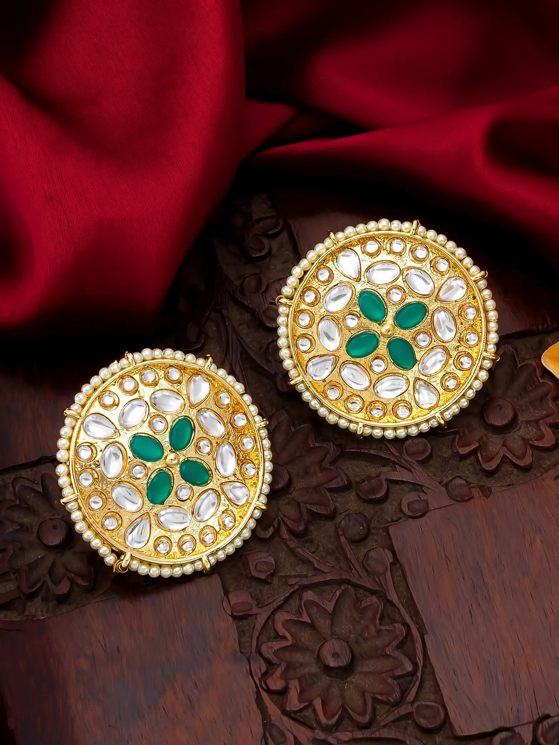 A pair of ornate Indian gold earrings decorated with white stones, green enamel, and a border of small white pearls, displayed on a carved wooden surface with a red satin cloth background.