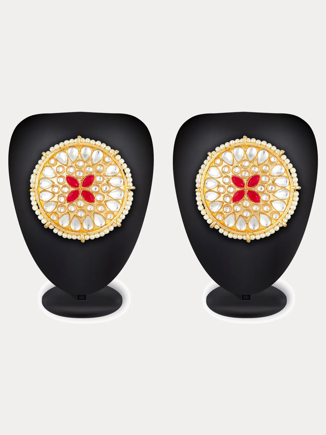 A pair of ornate gold-toned Indian jhumka earrings decorated with white stones and a red floral motif in the center, displayed on a carved wooden surface with red fabric.
