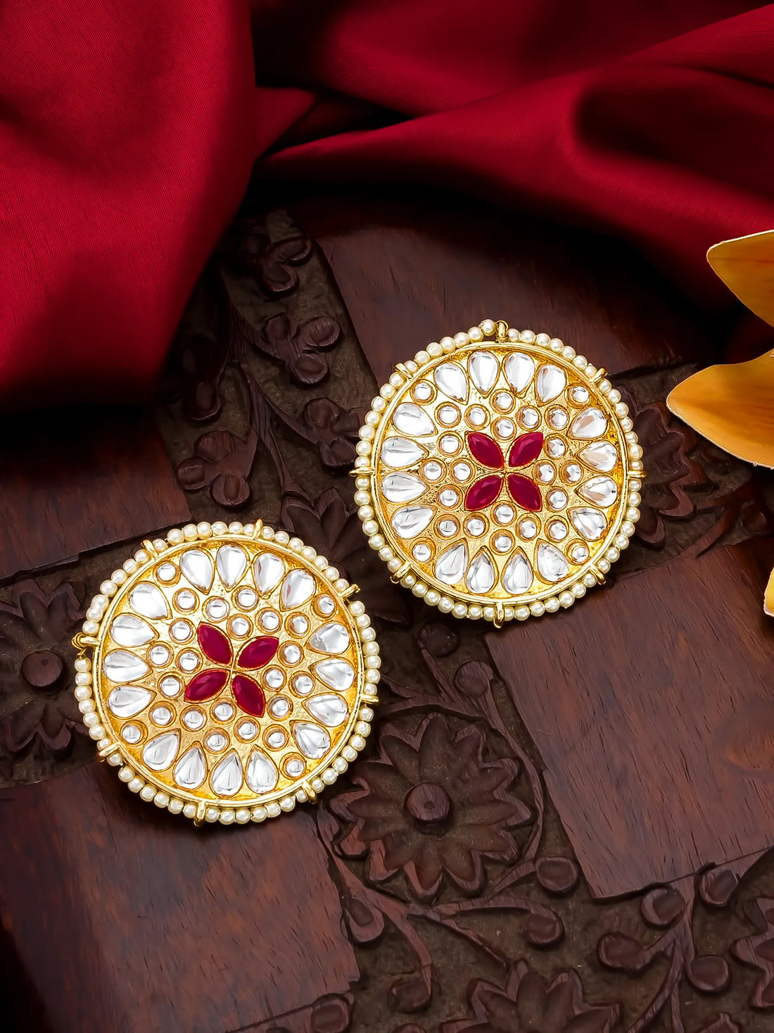 A pair of ornate gold-toned Indian jhumka earrings decorated with white stones and a red floral motif in the center, displayed on a carved wooden surface with red fabric.