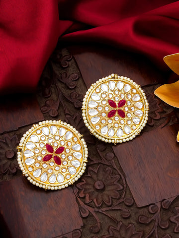 A pair of ornate gold-toned Indian jhumka earrings decorated with white stones and a red floral motif in the center, displayed on a carved wooden surface with red fabric.