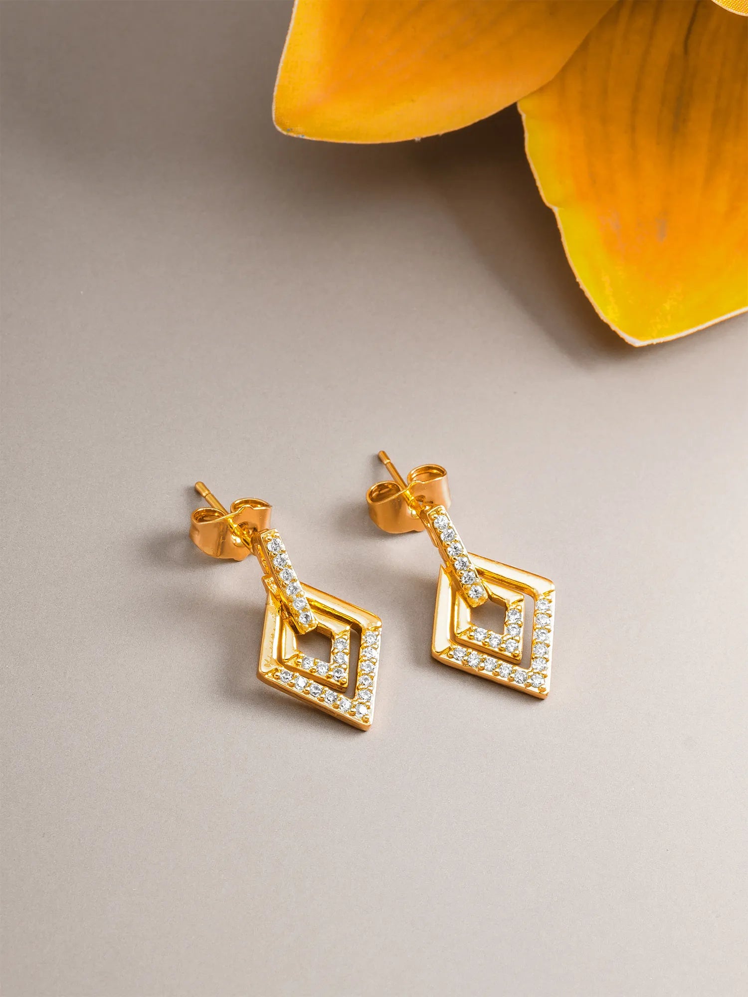 A pair of gold geometric drop earrings with clear stones, displayed on a neutral gray surface with a yellow flower petal in the background.