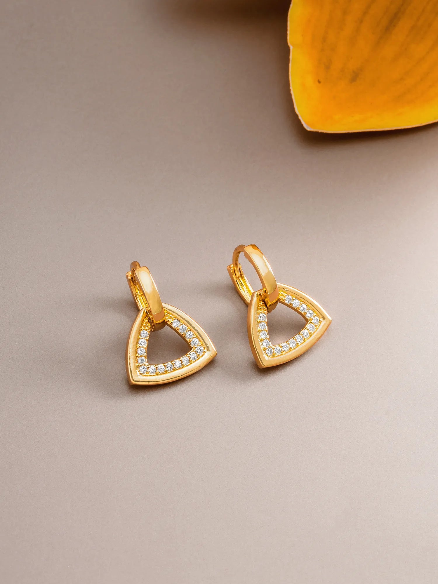 A pair of gold triangular hoop earrings with small clear stones along the front edges, displayed on a neutral taupe surface with a yellow leaf in the top right corner.