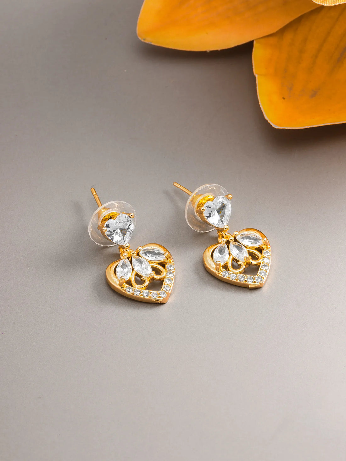 A pair of gold heart-shaped stud earrings with clear crystal drops, displayed on a neutral gray surface with a yellow flower in the background.