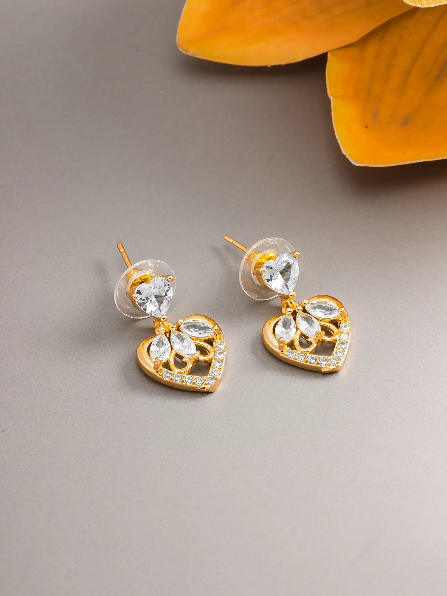 A pair of gold heart-shaped stud earrings with clear crystal drops, displayed on a neutral gray surface with a yellow flower in the background.