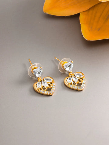 A pair of gold heart-shaped stud earrings with clear crystal drops, displayed on a neutral gray surface with a yellow flower in the background.