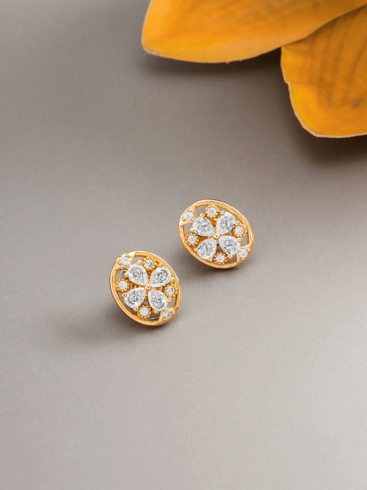 A pair of gold floral stud earrings with clear stones, displayed on a neutral gray surface with a yellow flower petal in the background.