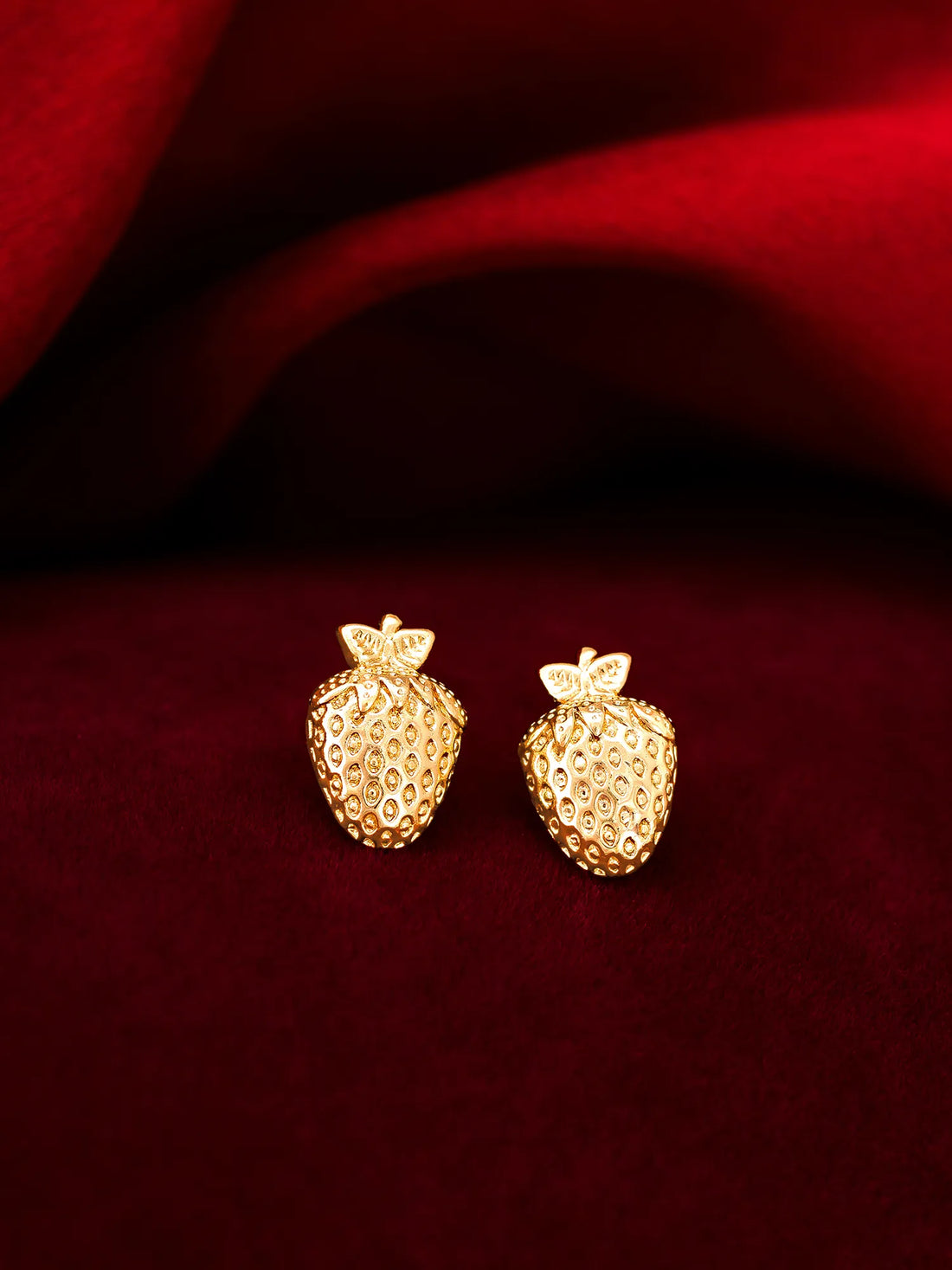 A pair of gold stud earrings shaped like textured strawberries, displayed on a dark red velvet surface with a matching red fabric backdrop.