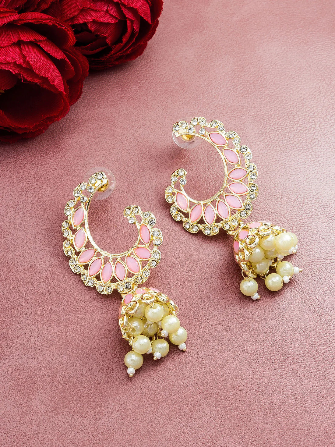 A pair of gold-plated crescent-shaped jhumka earrings with pink enamel details, clear rhinestones, and dangling cream pearl clusters, displayed on a textured pink surface with red roses in the corner.
