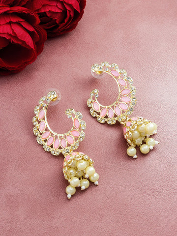 A pair of gold-plated crescent-shaped jhumka earrings with pink enamel details, clear rhinestones, and dangling cream pearl clusters, displayed on a textured pink surface with red roses in the corner.