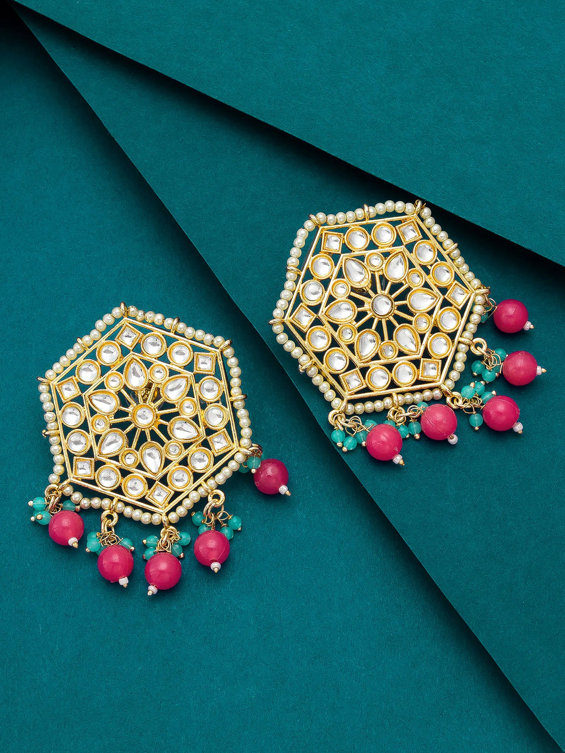 A pair of gold-toned geometric dangle earrings with white stones, green beads, and pink bead drops, arranged on a dark teal background.
