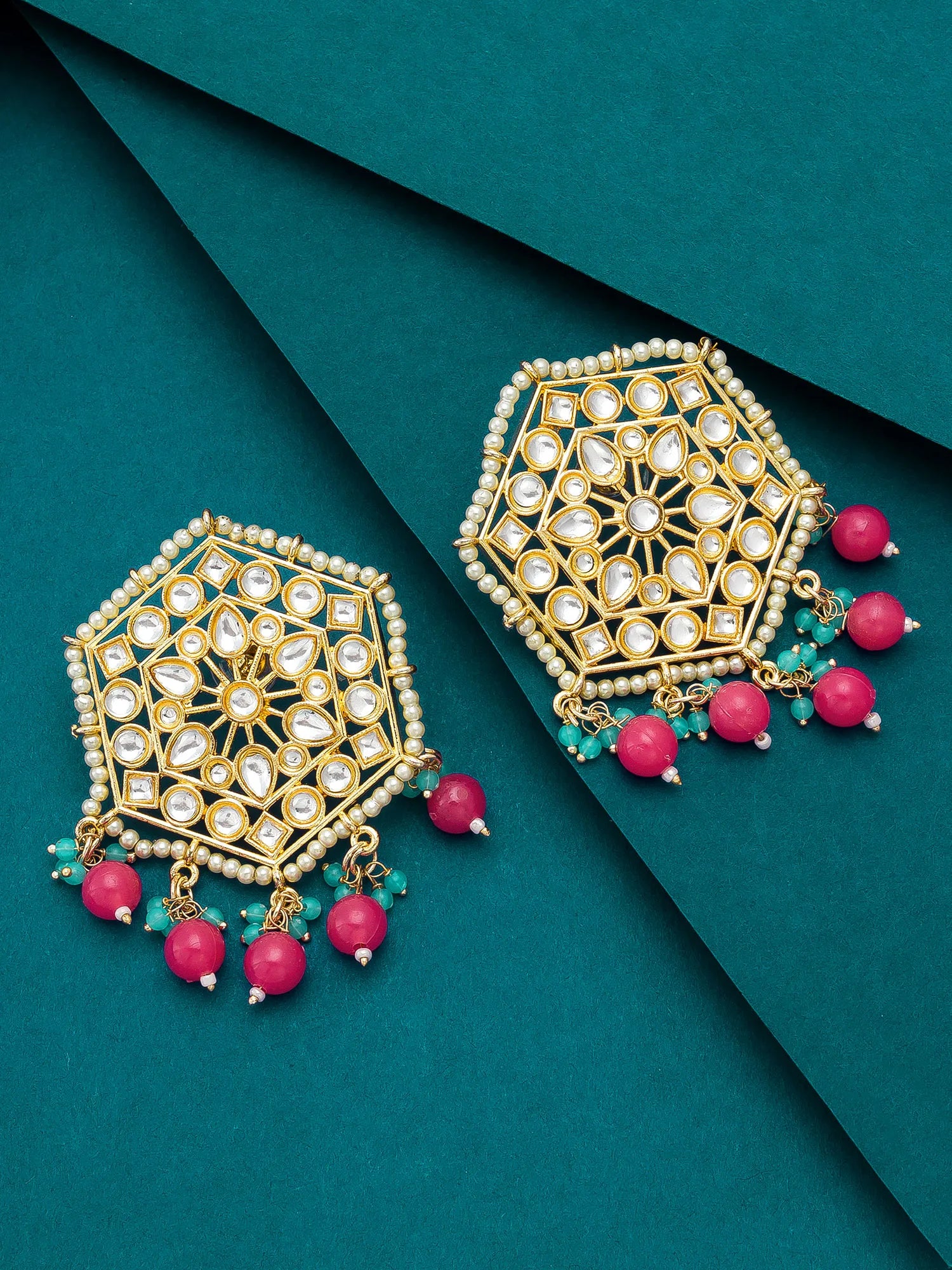 A pair of gold-toned geometric dangle earrings with white stones, green beads, and pink bead drops, arranged on a dark teal background.