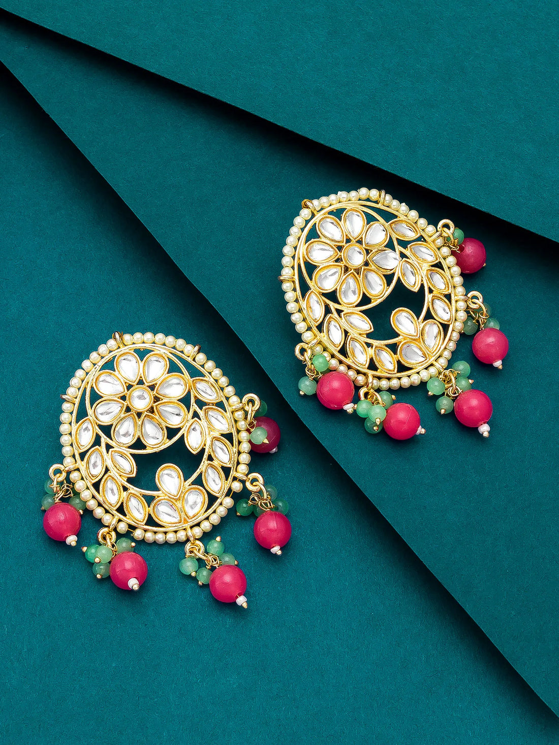 A pair of gold-toned Indian chandbali earrings with white inlay stones, pink bead drops, and green stone accents, displayed on a dark teal background.