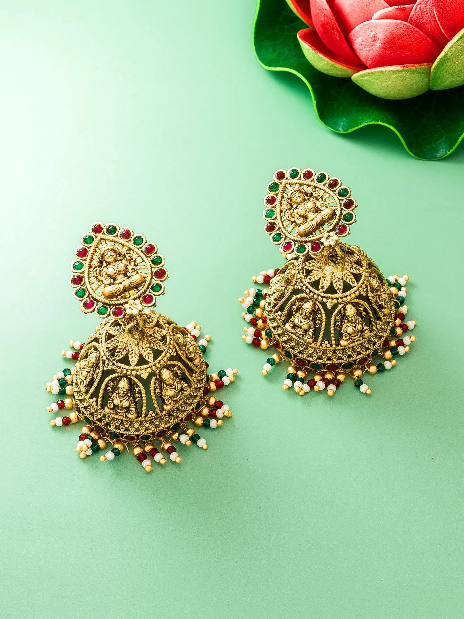 A pair of ornate gold-toned Indian jhumka earrings decorated with red and green stones and white bead tassels, displayed on a light green background with a decorative red lotus flower in the top right corner.