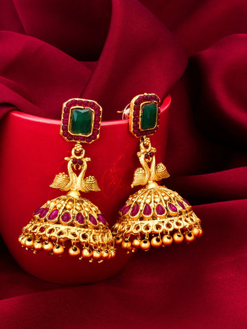 A pair of ornate gold jhumka earrings with green rectangular stones and purple stone accents, displayed on a red satin background.