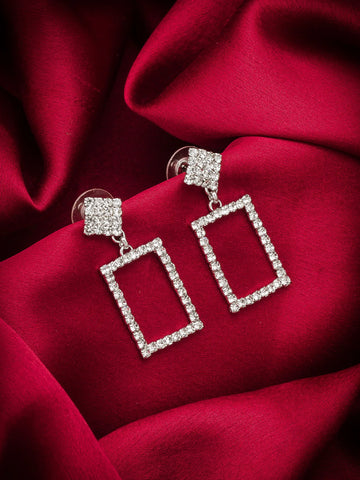 A pair of silver-tone drop earrings with square-shaped rectangular frames paved with clear cubic zirconia stones, resting on a draped deep red satin fabric background.