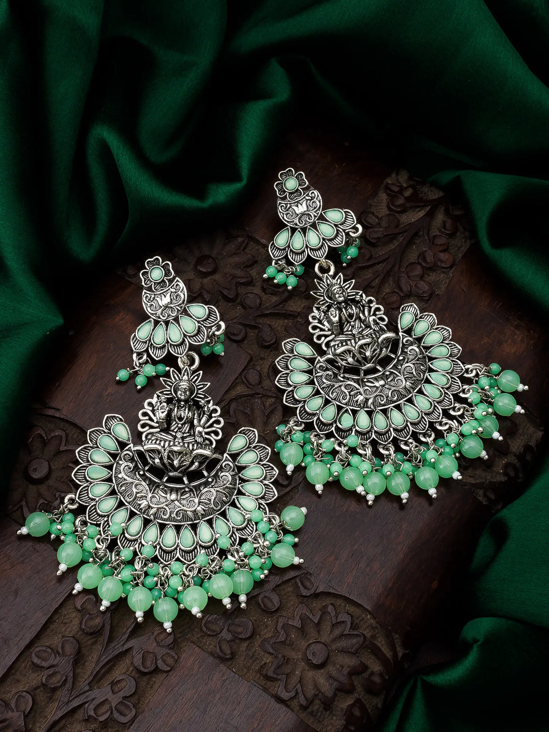 Ornate silver and green earrings on a dark surface with green fabric background