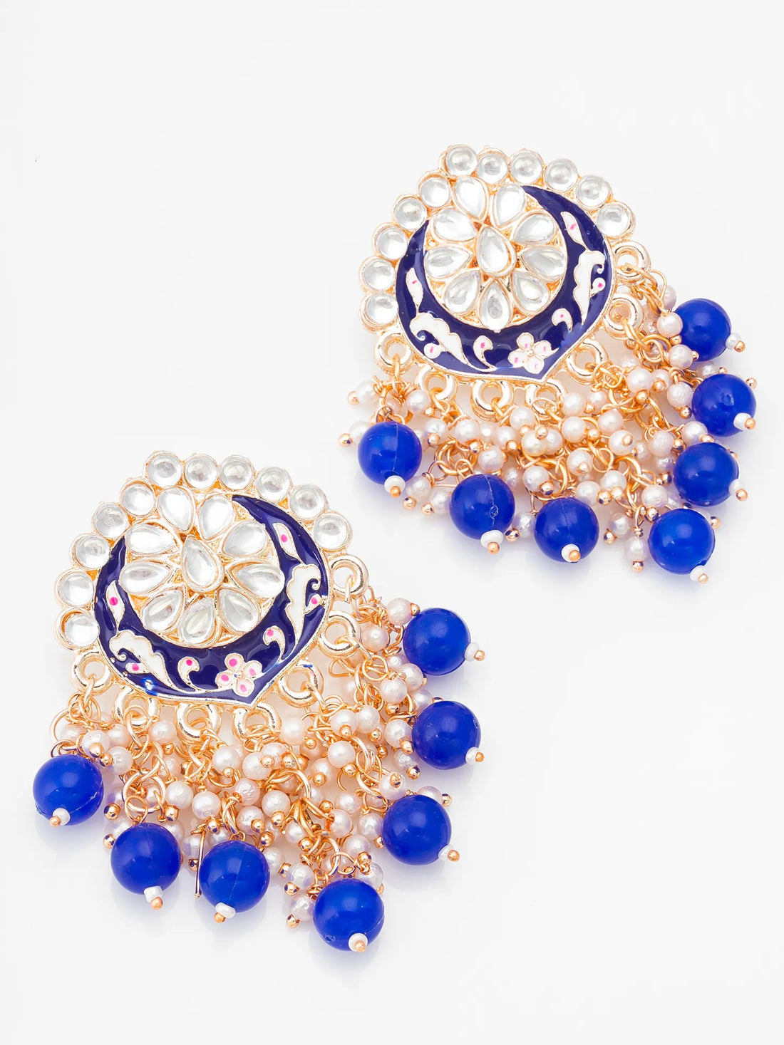 A pair of ornate gold-toned Indian jhumka earrings decorated with white faux pearls, blue enamel, and blue beads, displayed on a green surface with a wooden box and burlap cloth in the background.
