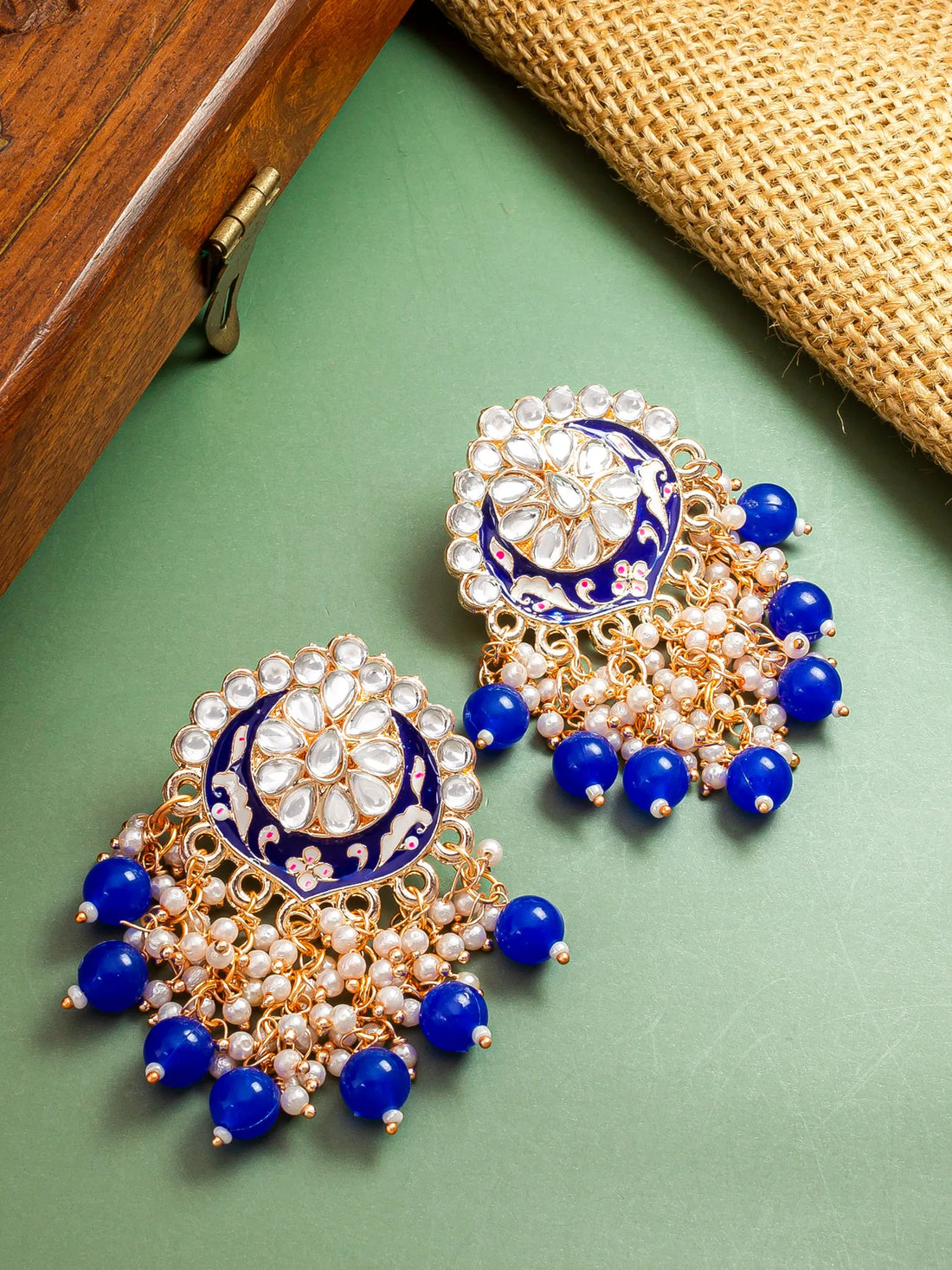 A pair of ornate gold-toned Indian jhumka earrings decorated with white faux pearls, blue enamel, and blue beads, displayed on a green surface with a wooden box and burlap cloth in the background.