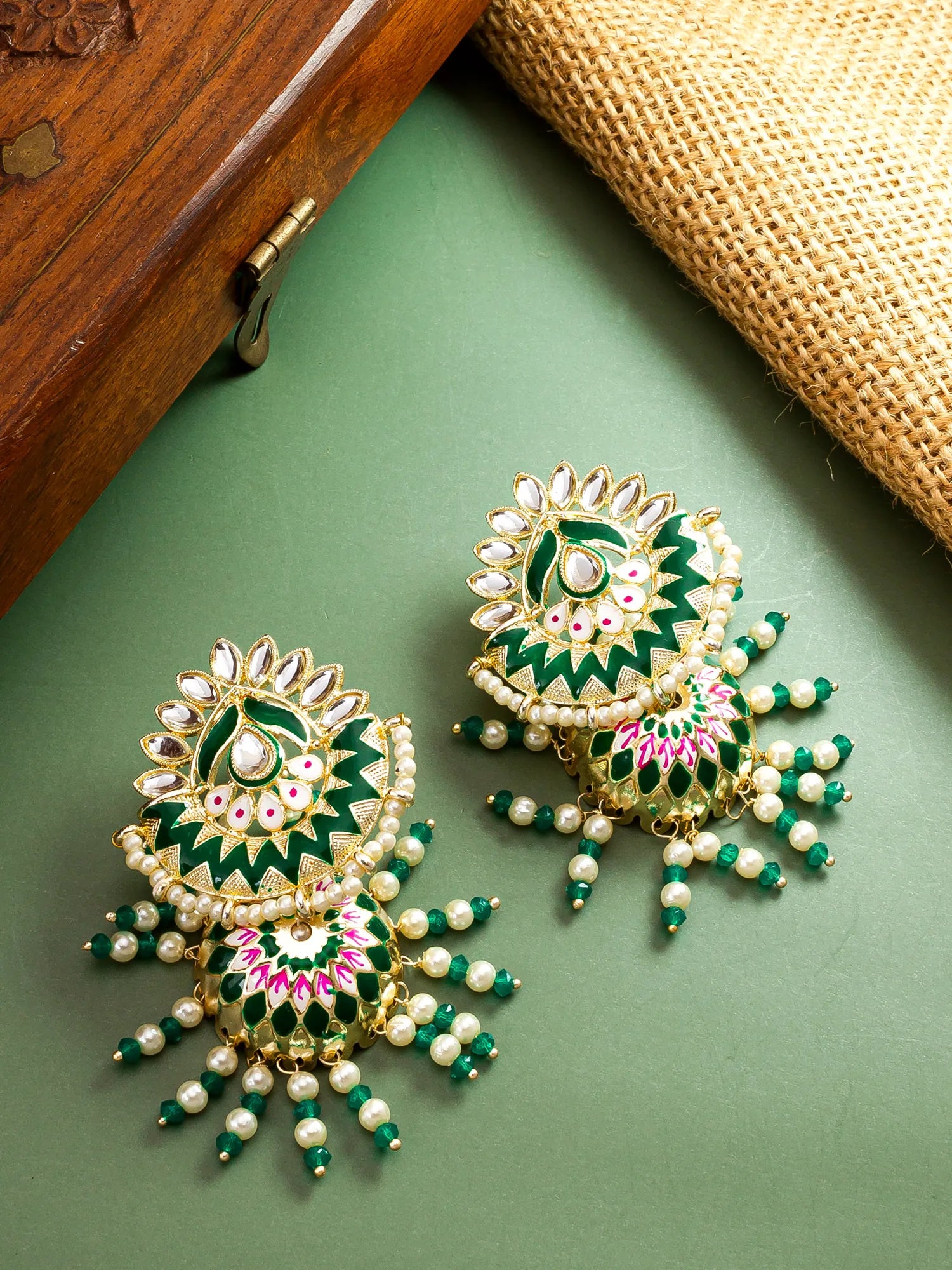 A pair of ornate gold-plated Indian jhumka earrings decorated with green and pink enamel, white faux pearls, and small green beads, displayed on a sage green surface with a wooden box and burlap cloth in the background.