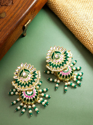 A pair of ornate gold-plated Indian jhumka earrings decorated with green and pink enamel, white faux pearls, and small green beads, displayed on a sage green surface with a wooden box and burlap cloth in the background.