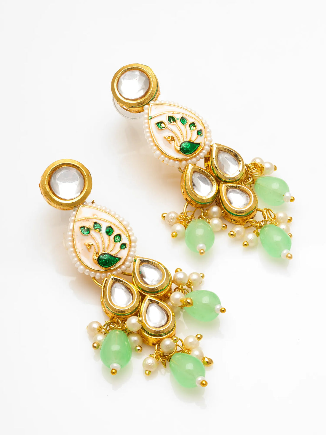 A pair of gold-plated Indian chandbali earrings decorated with white faux pearls, green stone accents, and light green teardrop beads, displayed on a pink surface with a burlap and maroon fabric background.