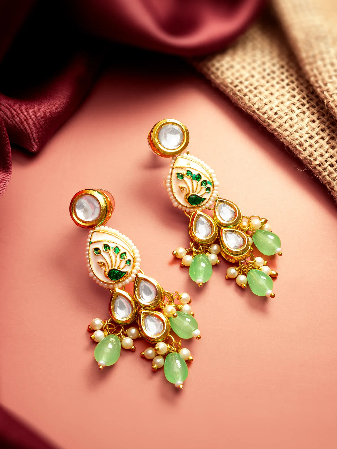 A pair of gold-plated Indian chandbali earrings decorated with white faux pearls, green stone accents, and light green teardrop beads, displayed on a pink surface with a burlap and maroon fabric background.