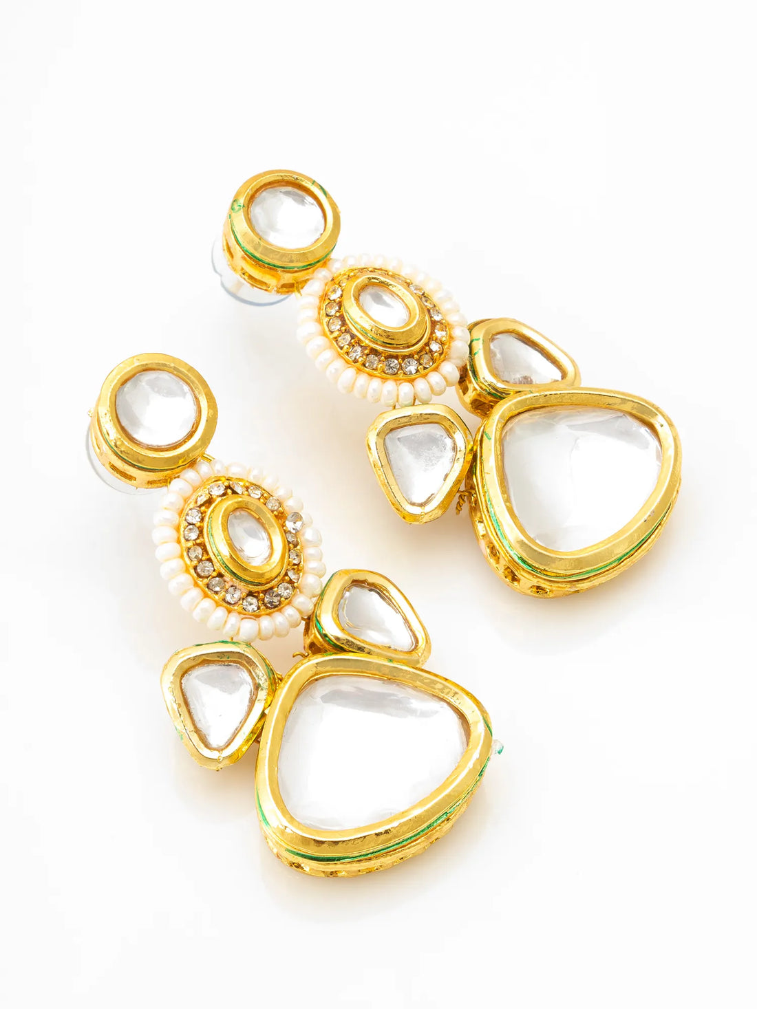 A pair of gold-plated kanzur earrings with white stones, displayed on a peach-colored surface with draped burgundy satin and a gold textured ribbon in the background.