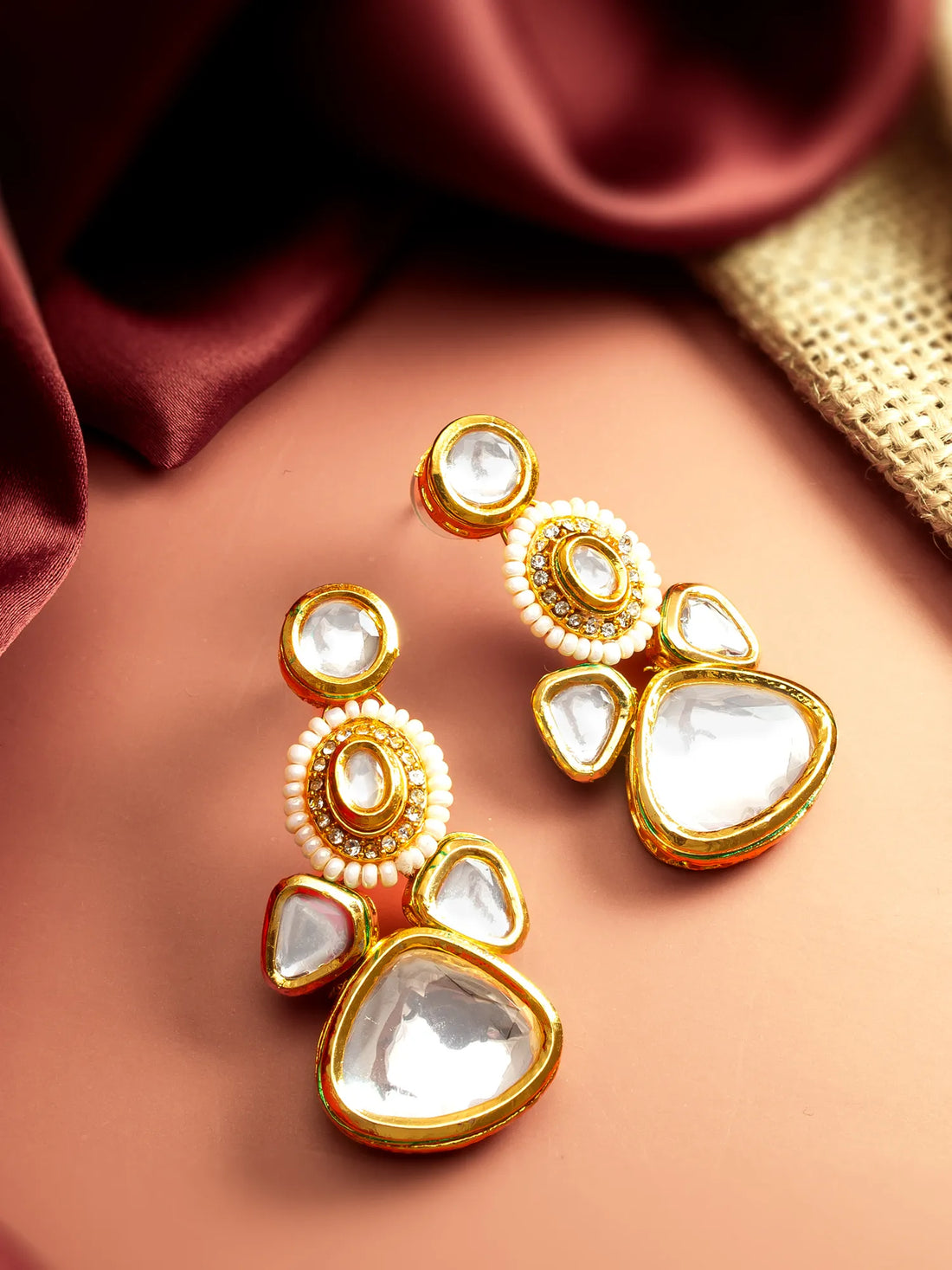A pair of gold-plated kanzur earrings with white stones, displayed on a peach-colored surface with draped burgundy satin and a gold textured ribbon in the background.