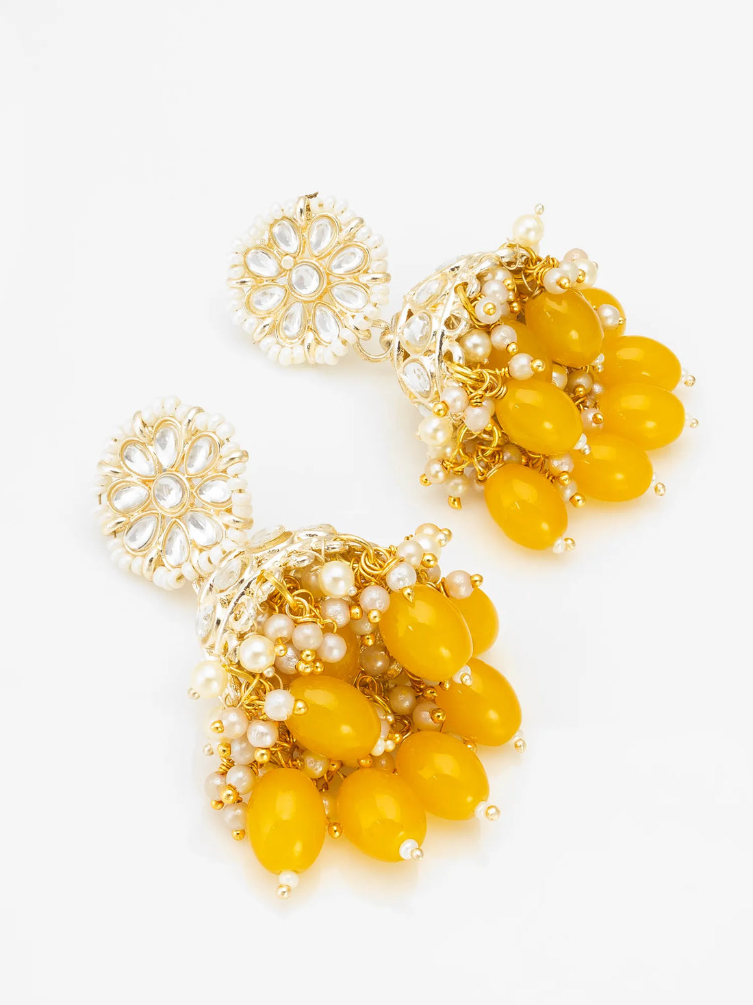 Gold-plated jhumka earrings with white stone floral tops and dangling amber-yellow beads, displayed against a dark brown carved wooden background.