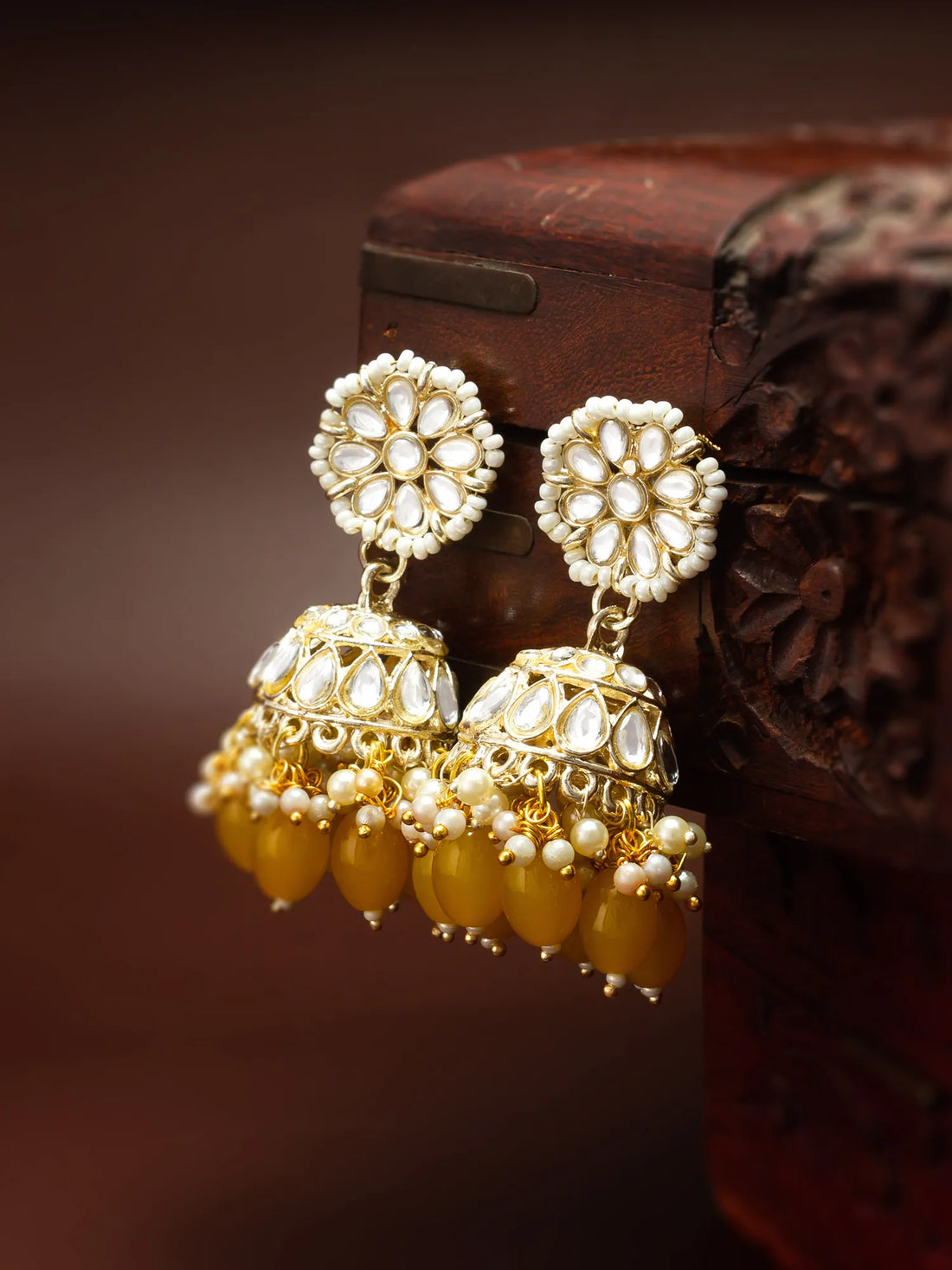 Gold-plated jhumka earrings with white stone floral tops and dangling amber-yellow beads, displayed against a dark brown carved wooden background.