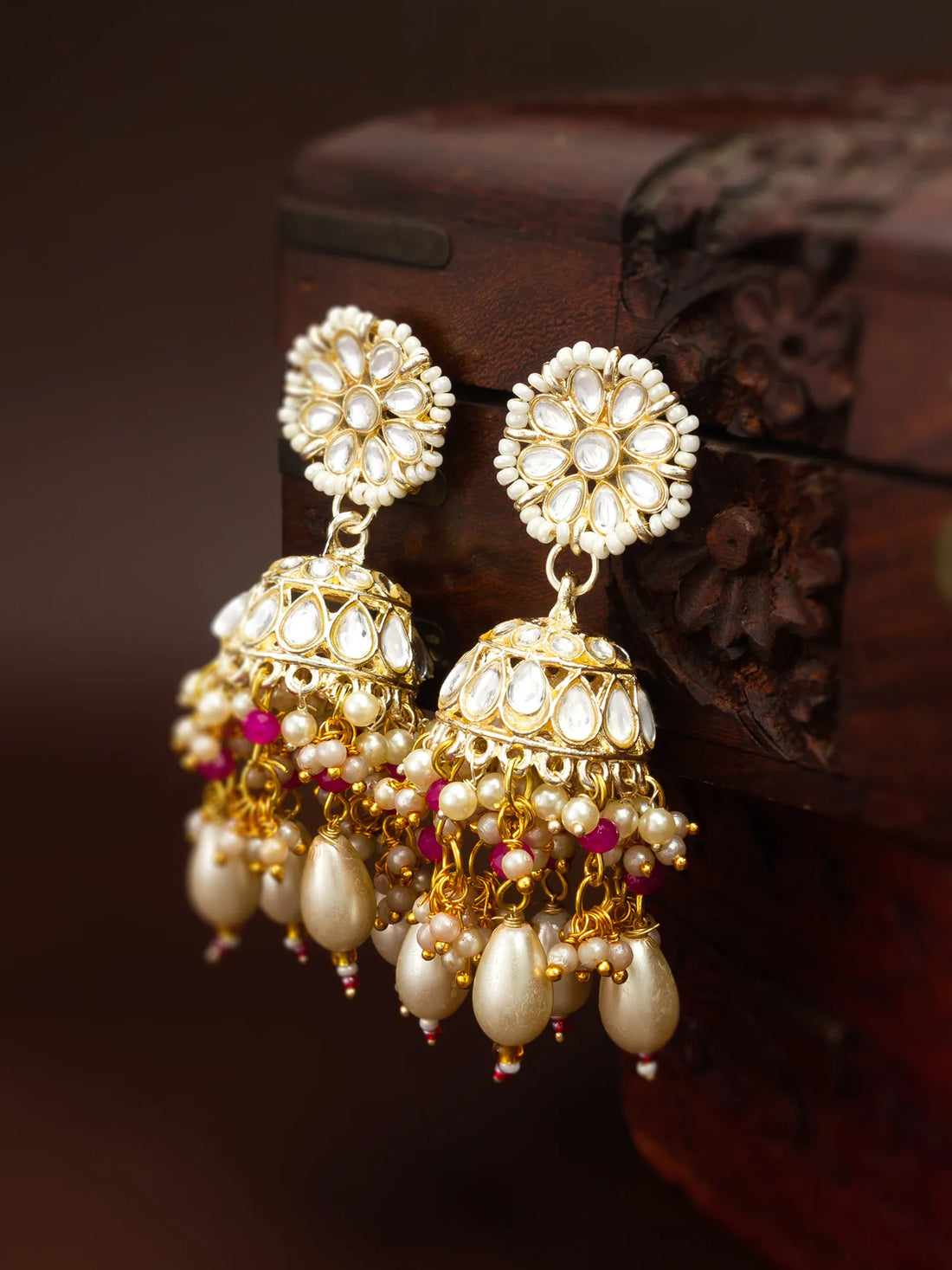 A pair of ornate gold and pearl Jhumka earrings with small pink bead accents, displayed against a dark carved wooden box.