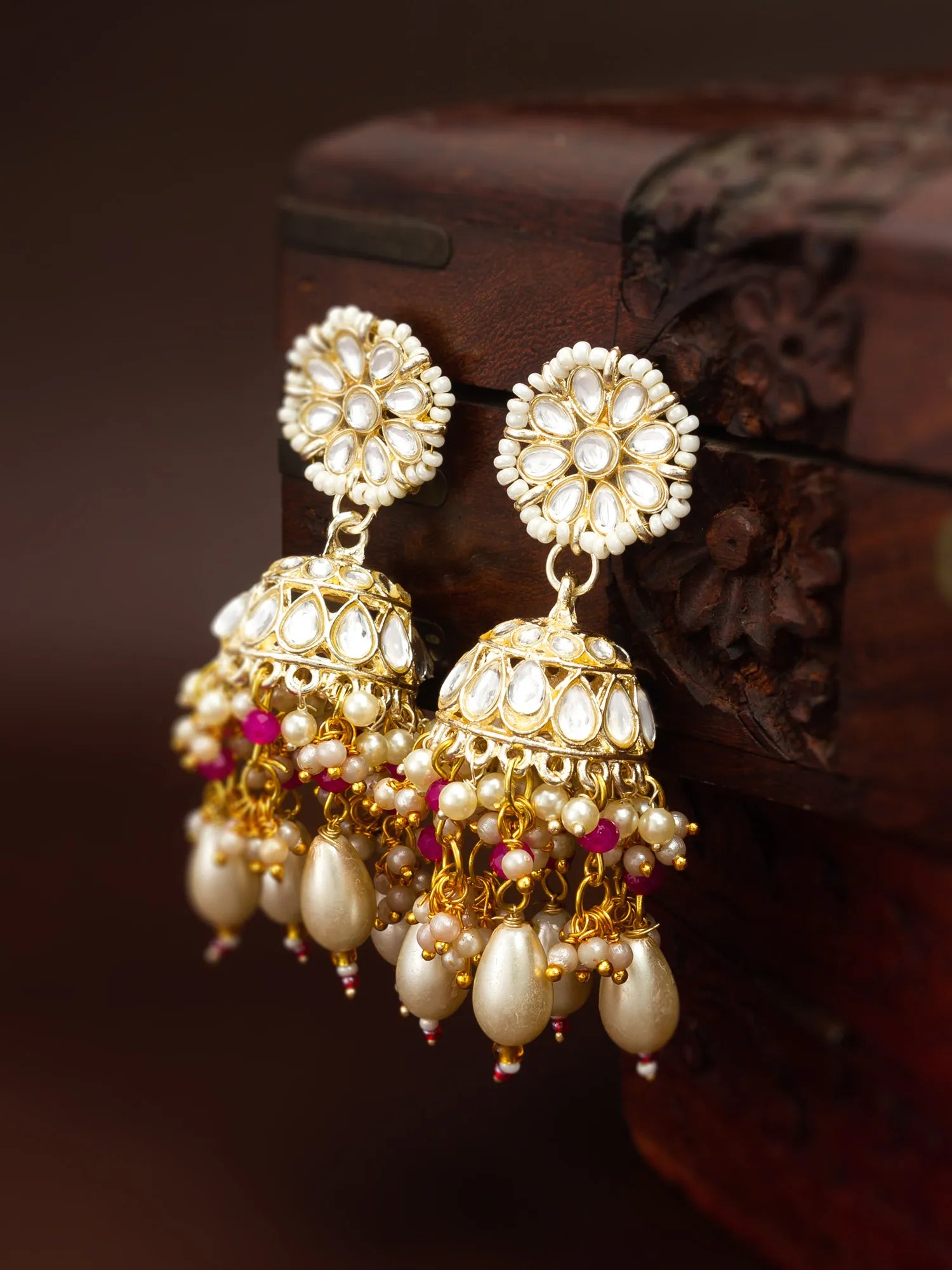 A pair of ornate gold and pearl Jhumka earrings with small pink bead accents, displayed against a dark carved wooden box.