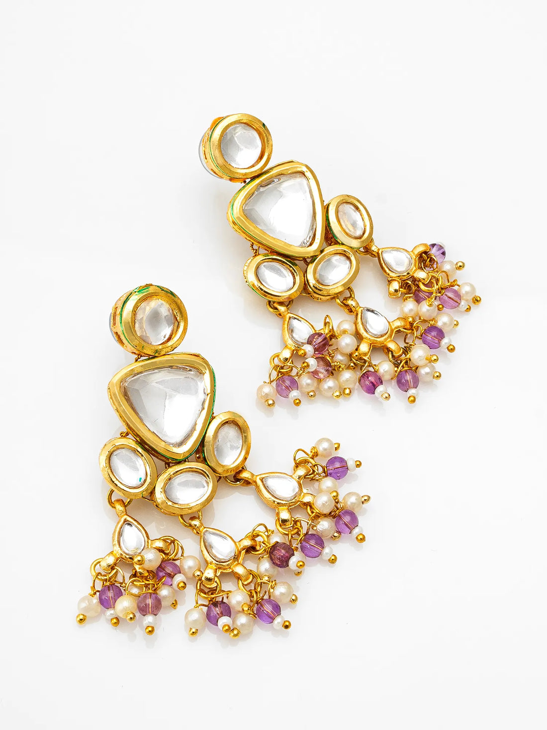 Gold-toned statement earrings with large white stones and dangling pearl-like beads with small purple accents, arranged on a pink surface with red fabric and burlap in the background.