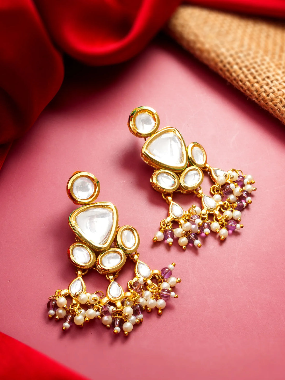 Gold-toned statement earrings with large white stones and dangling pearl-like beads with small purple accents, arranged on a pink surface with red fabric and burlap in the background.