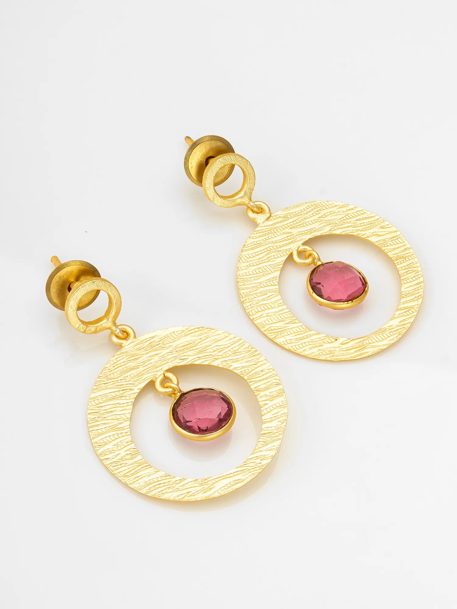 A pair of gold-toned dangle earrings with a textured circular disc, a small hanging loop, and a pink gemstone accent, shown on a plain white background.