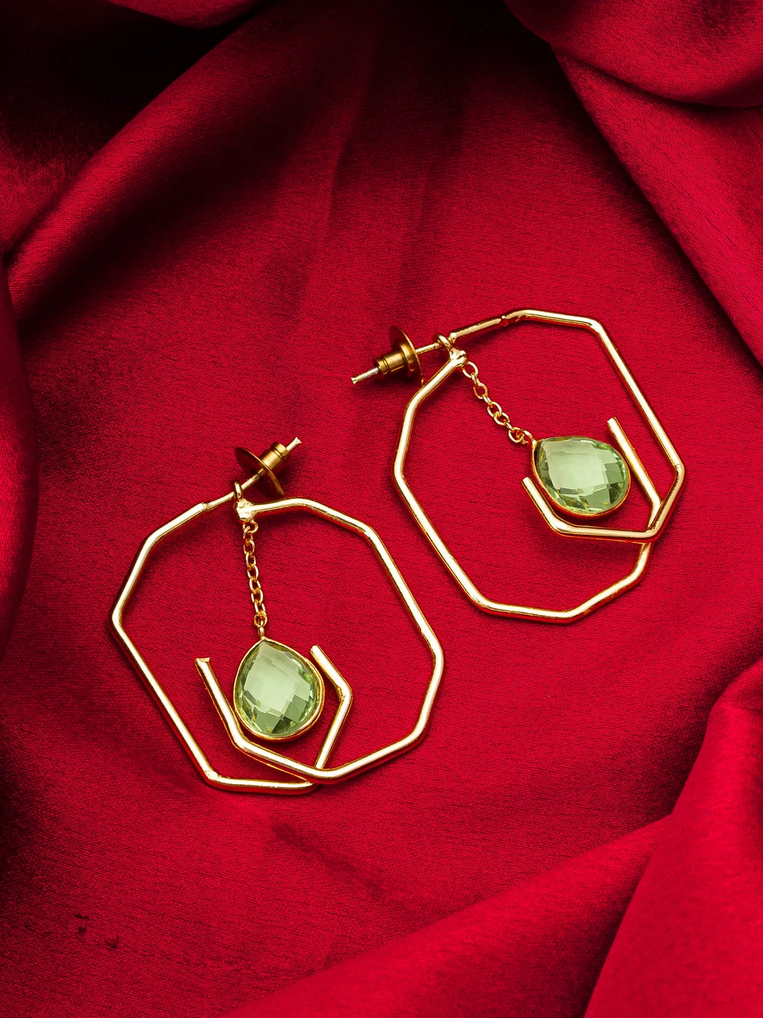Gold geometric hoop earrings with dangling light green faceted gemstones, displayed on a draped red fabric background.