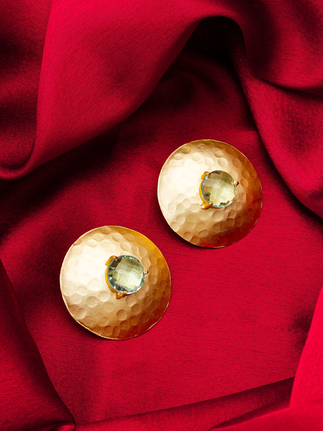Two hammered gold-tone oval earrings with central clear gemstones, arranged on draped red fabric.