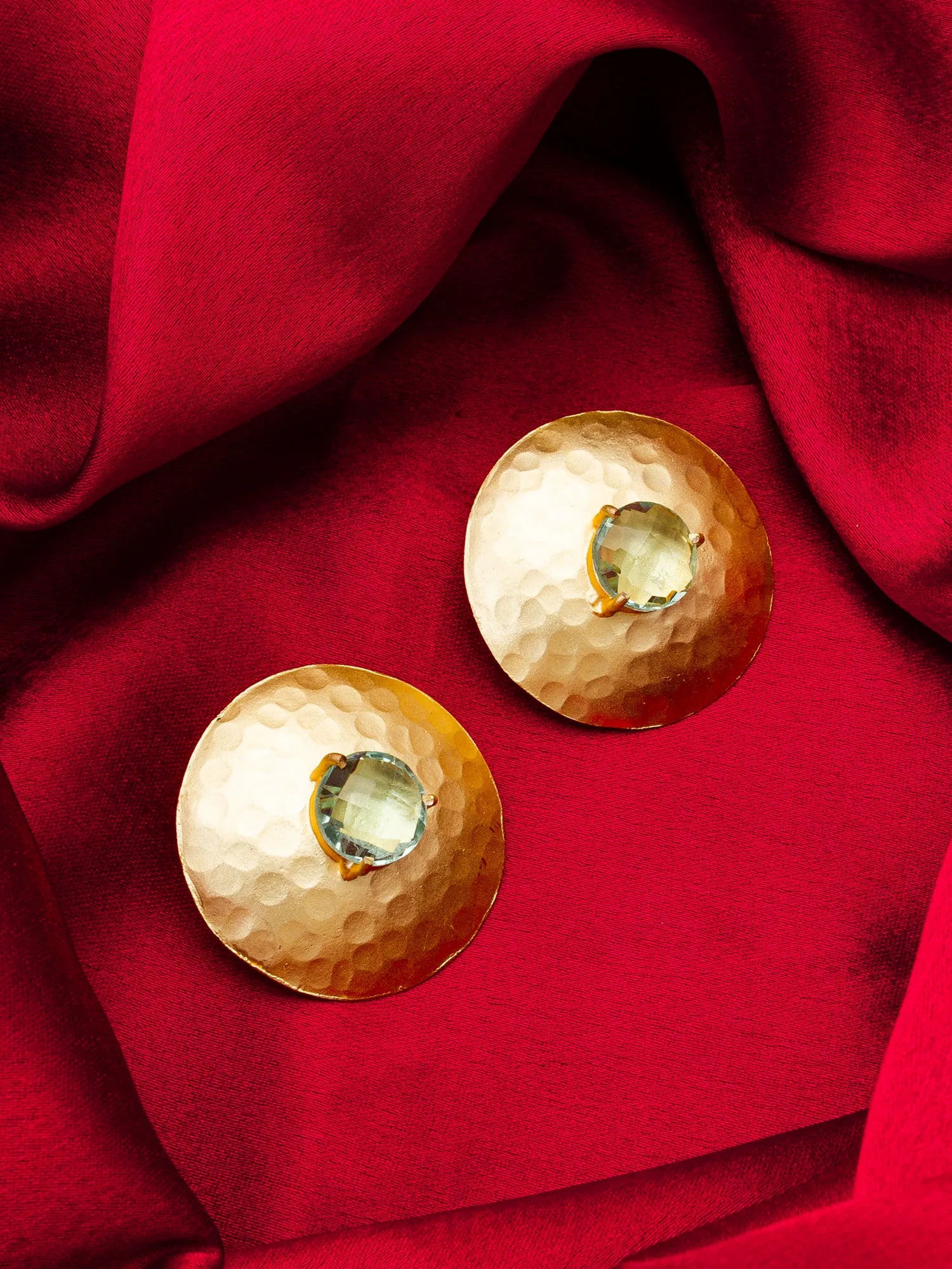 Two hammered gold-tone oval earrings with central clear gemstones, arranged on draped red fabric.