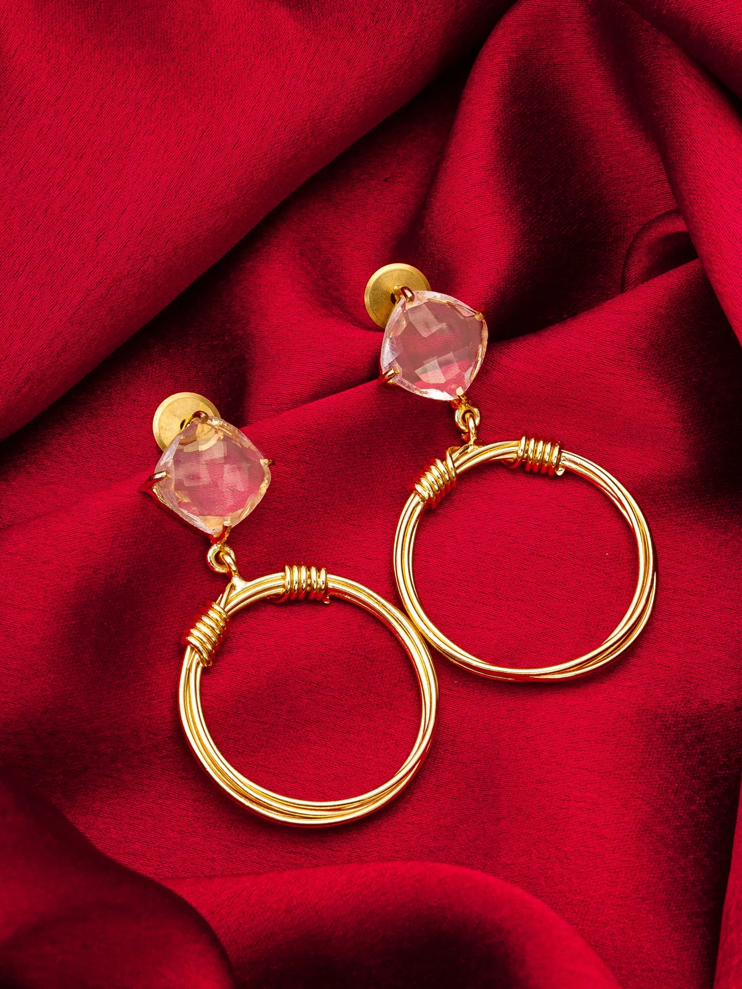 Gold hoop earrings with pink heart-shaped stones on a red fabric background