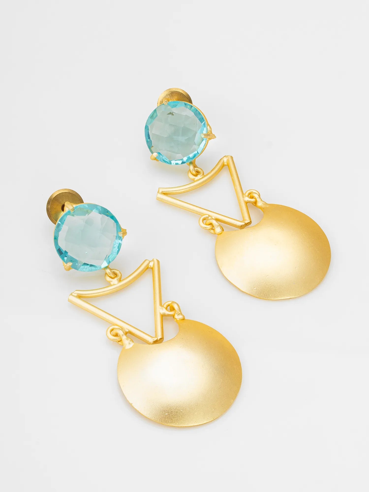 A pair of gold statement earrings featuring a round light blue gemstone and a large textured gold disc dangle, photographed on a clean white background.