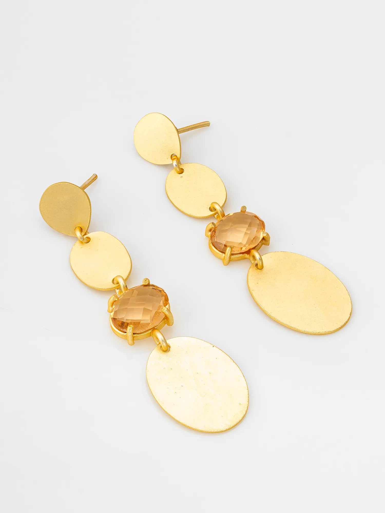 A pair of gold-toned dangle earrings featuring flat oval discs, faceted amber-orange gemstone accents, and larger textured oval drops, displayed on a clean white background.