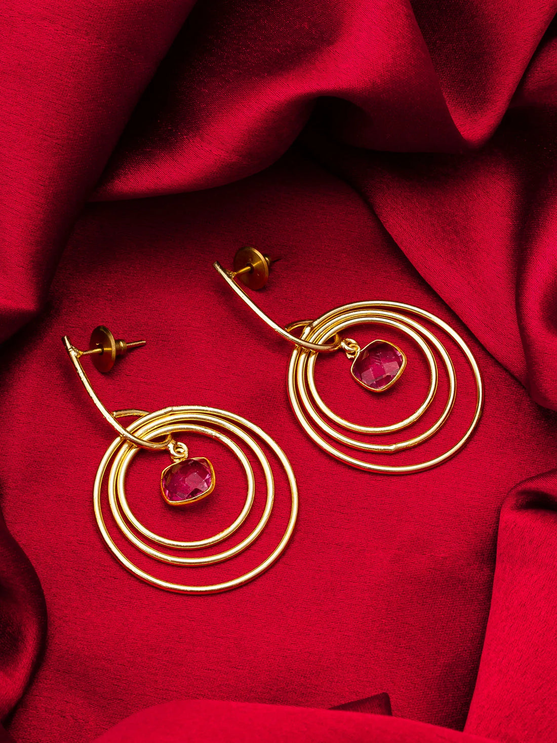 Gold hoop earrings with red gemstones on a red fabric background