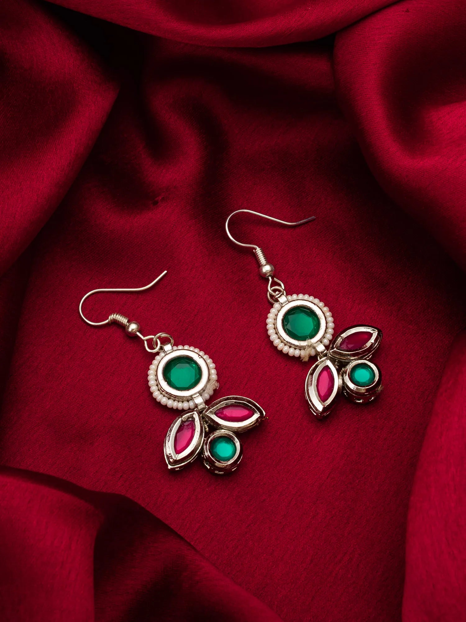 A pair of silver-toned traditional Indian earrings with green and pink stones, displayed on a draped red satin fabric background.